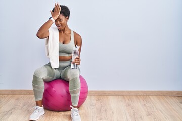 African american woman wearing sportswear sitting on pilates ball surprised with hand on head for...