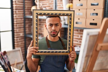Young hispanic man sitting at art studio with empty frame skeptic and nervous, frowning upset because of problem. negative person.