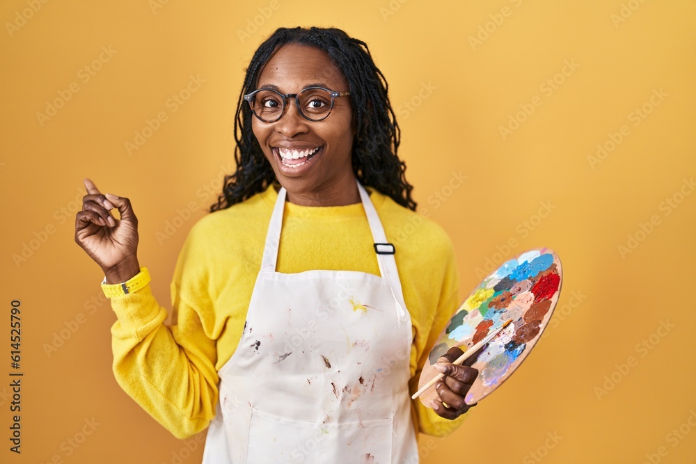 Sticker african woman holding painter palette with a big smile on face, pointing with hand finger to the sid