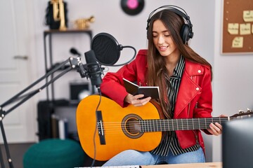 Fototapeta na wymiar Young beautiful hispanic woman musician playing classical guitar reading notebook at music studio