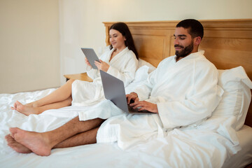 Obraz premium Husband And Wife Browsing Internet On Computers Lying In Bedroom