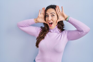 Fototapeta premium Young brunette woman standing over blue background smiling cheerful playing peek a boo with hands showing face. surprised and exited