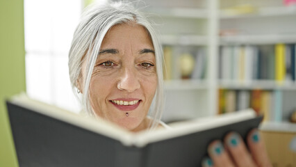 Middle age grey-haired woman student reading book smiling at library university