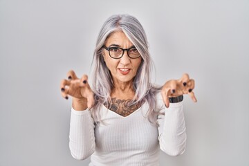 Middle age woman with grey hair standing over white background smiling funny doing claw gesture as...