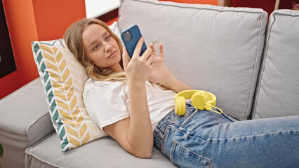 Young blonde woman using smartphone lying on the sofa at home