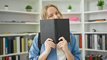 Young blonde woman student standing covering face with book at library university