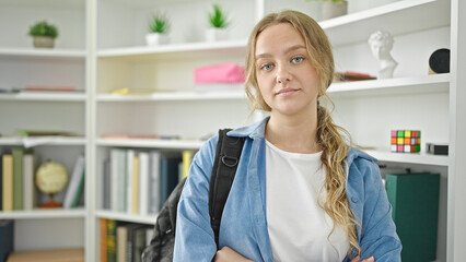 Young blonde woman student wearing backpack with relaxed expression and arms crossed gesture at library university