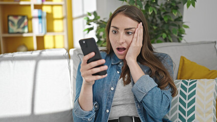 Young beautiful hispanic woman using smartphone with surprise expression at home