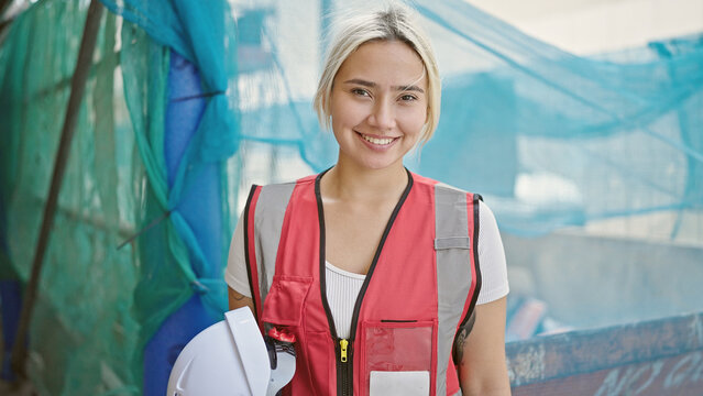 Young beautiful hispanic woman architect smiling confident holding hardhat at street