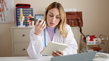 Young beautiful hispanic woman doctor sending voice message by smartphone reading notebook at clinic