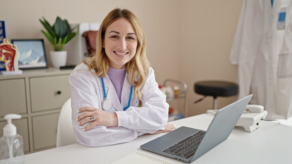 Young beautiful hispanic woman doctor using laptop working sitting on table with arms crossed gesture at clinic