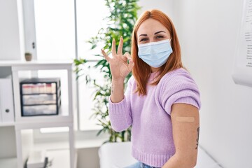 Young woman getting vaccine showing arm with band aid doing ok sign with fingers, smiling friendly gesturing excellent symbol