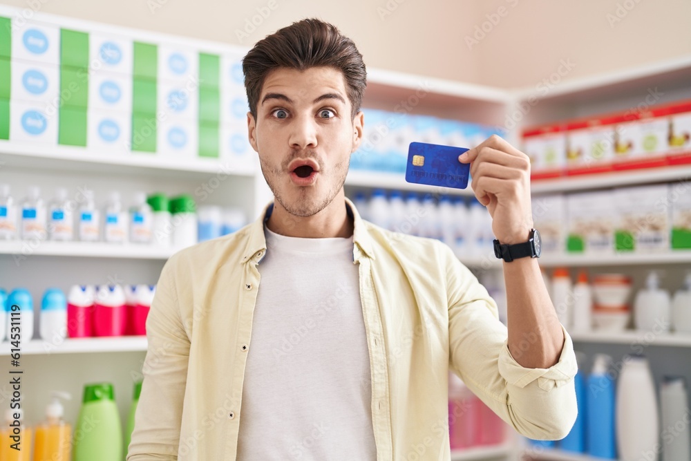 Sticker young hispanic man at pharmacy drugstore holding credit card scared and amazed with open mouth for s