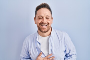 Middle age caucasian man standing over blue background smiling and laughing hard out loud because funny crazy joke with hands on body.