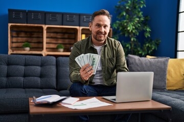 Young caucasian man using laptop counting dollars at home