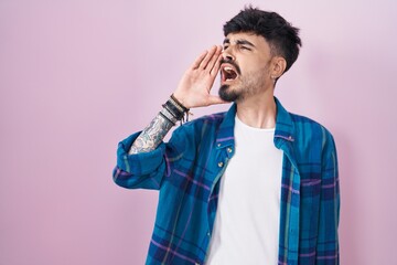 Young hispanic man with beard standing over pink background shouting and screaming loud to side with hand on mouth. communication concept.