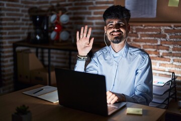 Young hispanic man with beard working at the office at night showing and pointing up with fingers number five while smiling confident and happy.