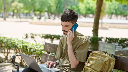 Young hispanic man tourist using laptop talking on smartphone at park