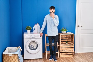 Young hispanic man with beard doing laundry standing at laundry room covering mouth with hand, shocked and afraid for mistake. surprised expression