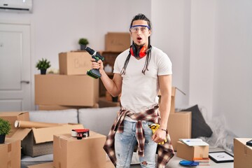 Hispanic man with long hair holding screwdriver at new home scared and amazed with open mouth for surprise, disbelief face