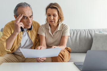 Household papers. Serious retired spouses using laptop and reading bills, looking through...