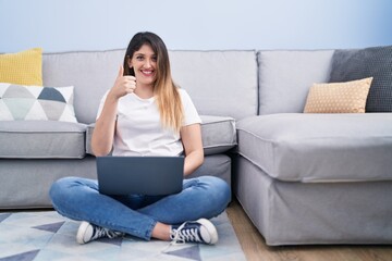 Obraz premium Young brunette woman sitting on the floor at home using laptop smiling happy and positive, thumb up doing excellent and approval sign