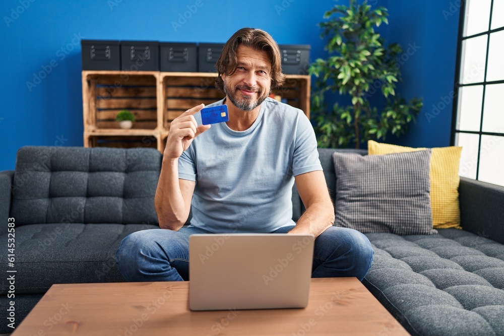 Sticker middle age man using laptop and credit card sitting on sofa at home