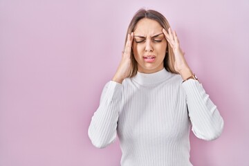 Young blonde woman wearing white sweater over pink background with hand on head for pain in head because stress. suffering migraine.