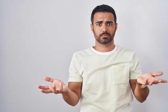 Hispanic man with beard standing over isolated background clueless and confused with open arms, no idea concept.