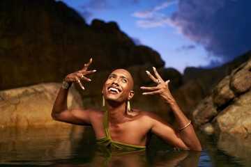 Smiling gay black lgbtq man poses with jewelry in water inside natural rocky pond at night....
