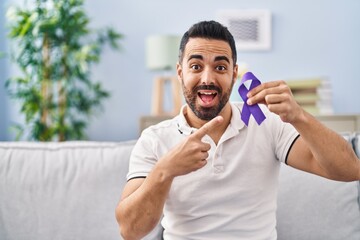 Young hispanic man with beard holding purple ribbon awareness smiling happy pointing with hand and finger