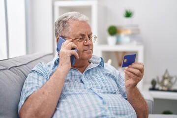 Middle age grey-haired man talking on smartphone holding credit card sitting on sofa at home