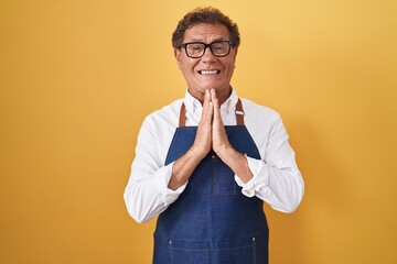Middle age hispanic man wearing professional cook apron praying with hands together asking for...