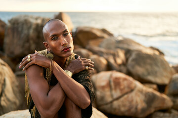 Non-binary black person in luxury dress on rocks in ocean . Trans ethnic fashion model wearing...