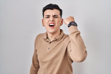 Non binary person standing over isolated background angry and mad raising fist frustrated and furious while shouting with anger. rage and aggressive concept.