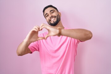 Hispanic young man standing over pink background smiling in love doing heart symbol shape with hands. romantic concept.