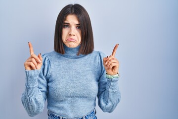 Young hispanic woman standing over blue background pointing up looking sad and upset, indicating direction with fingers, unhappy and depressed.