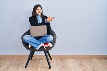 Young hispanic woman sitting on chair using computer laptop pointing aside with hands open palms showing copy space, presenting advertisement smiling excited happy