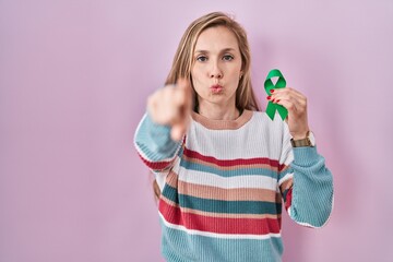 Young blonde woman holding support green ribbon pointing with finger to the camera and to you, confident gesture looking serious