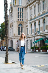 Smiling woman wallking on historic street Batumi city in Georgia. Young woman wearing jacket and jeans holding bag. In front of architectural buildings. Vertical composition. 