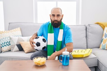 Young hispanic man with beard and tattoos football hooligan holding ball supporting team thinking...