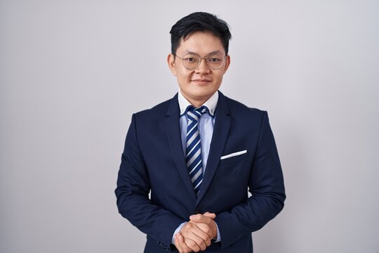 Young Asian Man Wearing Business Suit And Tie With Hands Together And Crossed Fingers Smiling Relaxed And Cheerful. Success And Optimistic