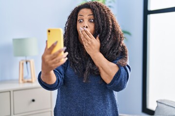 Plus size hispanic woman doing video call with smartphone covering mouth with hand, shocked and afraid for mistake. surprised expression