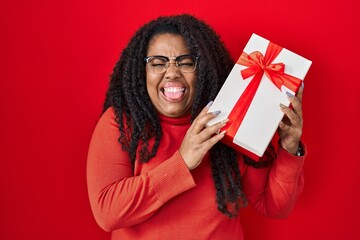 Plus size hispanic woman holding gift sticking tongue out happy with funny expression.