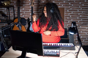 Plus size hispanic woman playing piano at music studio smiling with happy face looking and pointing to the side with thumb up.