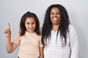 Mother and young daughter standing over white background showing and pointing up with finger number one while smiling confident and happy.