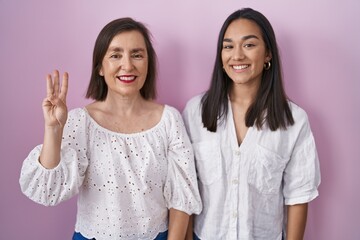 Hispanic mother and daughter together showing and pointing up with fingers number three while smiling confident and happy.