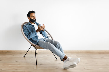 Arab guy drinking hot coffee in armchair against white wall