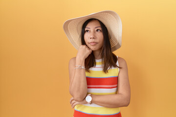 Middle age chinese woman wearing summer hat over yellow background with hand on chin thinking about question, pensive expression. smiling with thoughtful face. doubt concept.