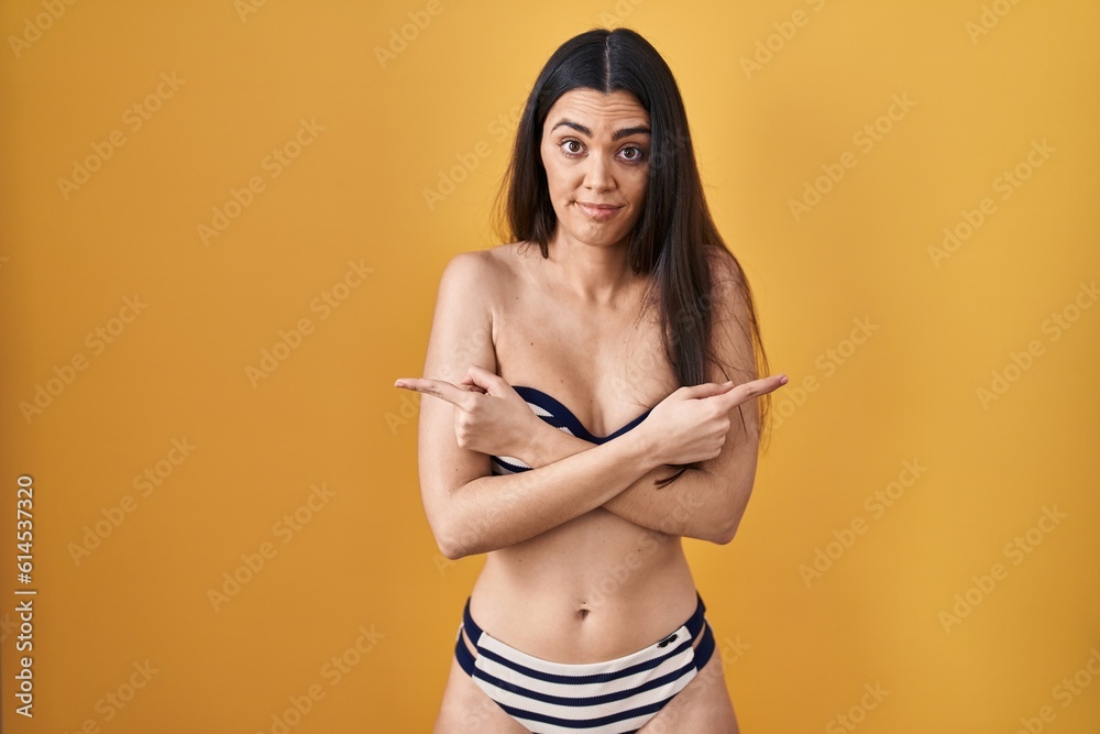 Wall mural young brunette woman wearing bikini over yellow background pointing to both sides with fingers, diff